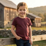 Young girl in a maroon shirt standing by a wooden fence with a barn and mountains in the background.