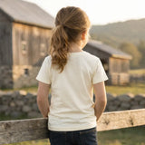 Child standing by a wooden fence looking at a barn in a rural setting