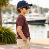 Young boy wearing a navy cap with an American flag, sunglasses, and a maroon shirt by a waterfront.