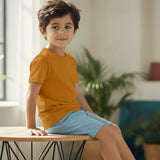 Child wearing an orange shirt and blue shorts sitting on a chair indoors.